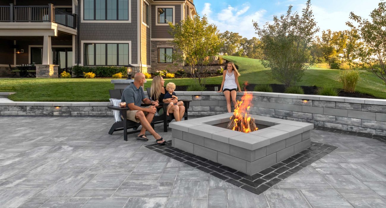 Family Sitting on Unilock Patio by Fire Pit and Person Sitting on Surrounding Retaining Wall